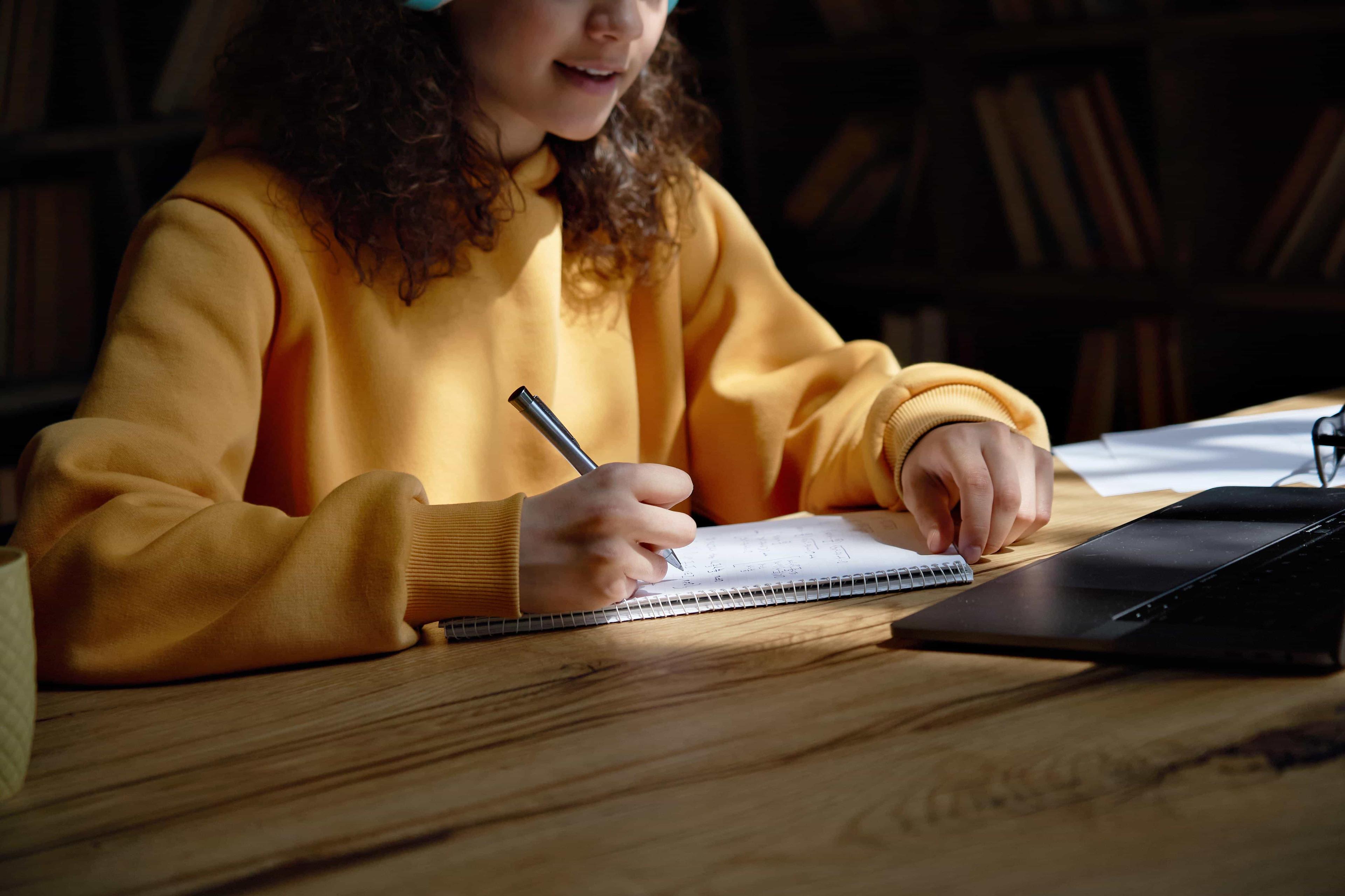 A GCSE English student sitting at a table with a pen and notebook, engaged in creative writing.