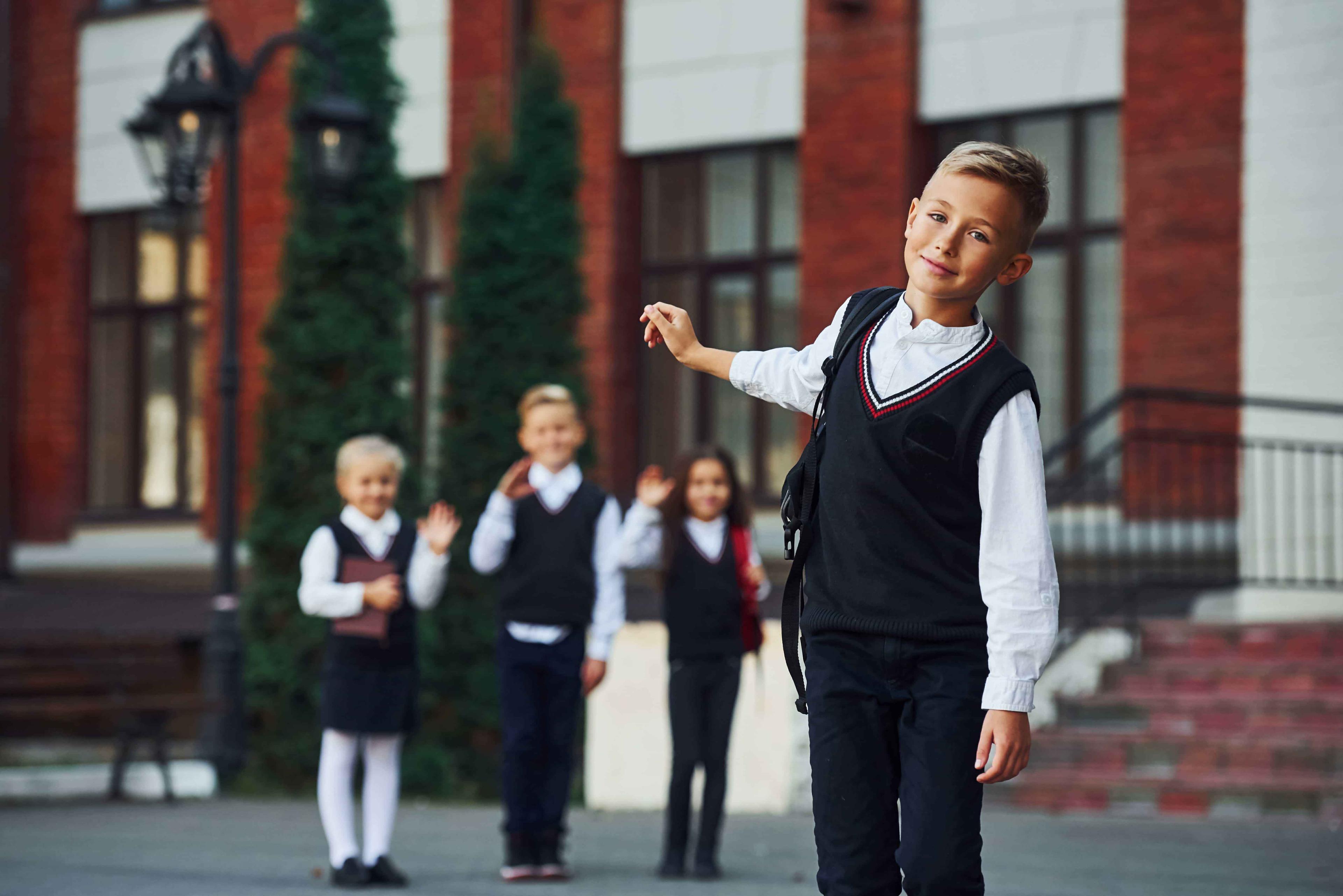 A preparatory school student tilts his head, hand raised, with blurred friends in the background in front of the school..jpeg