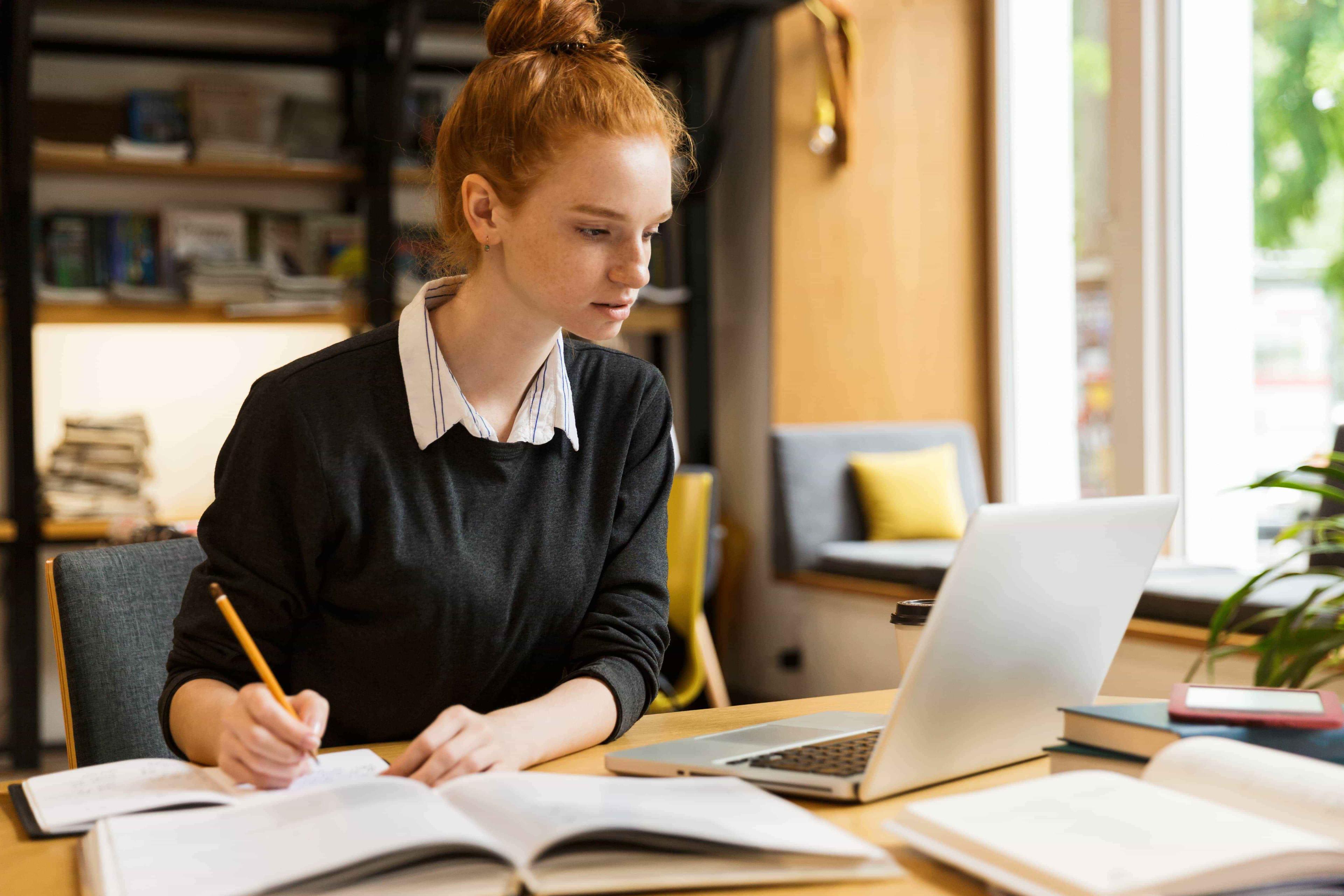 An IB student gazes at her laptop screen while jotting down notes in her notebook, engaged in her 1-1 online tutoring lesson.