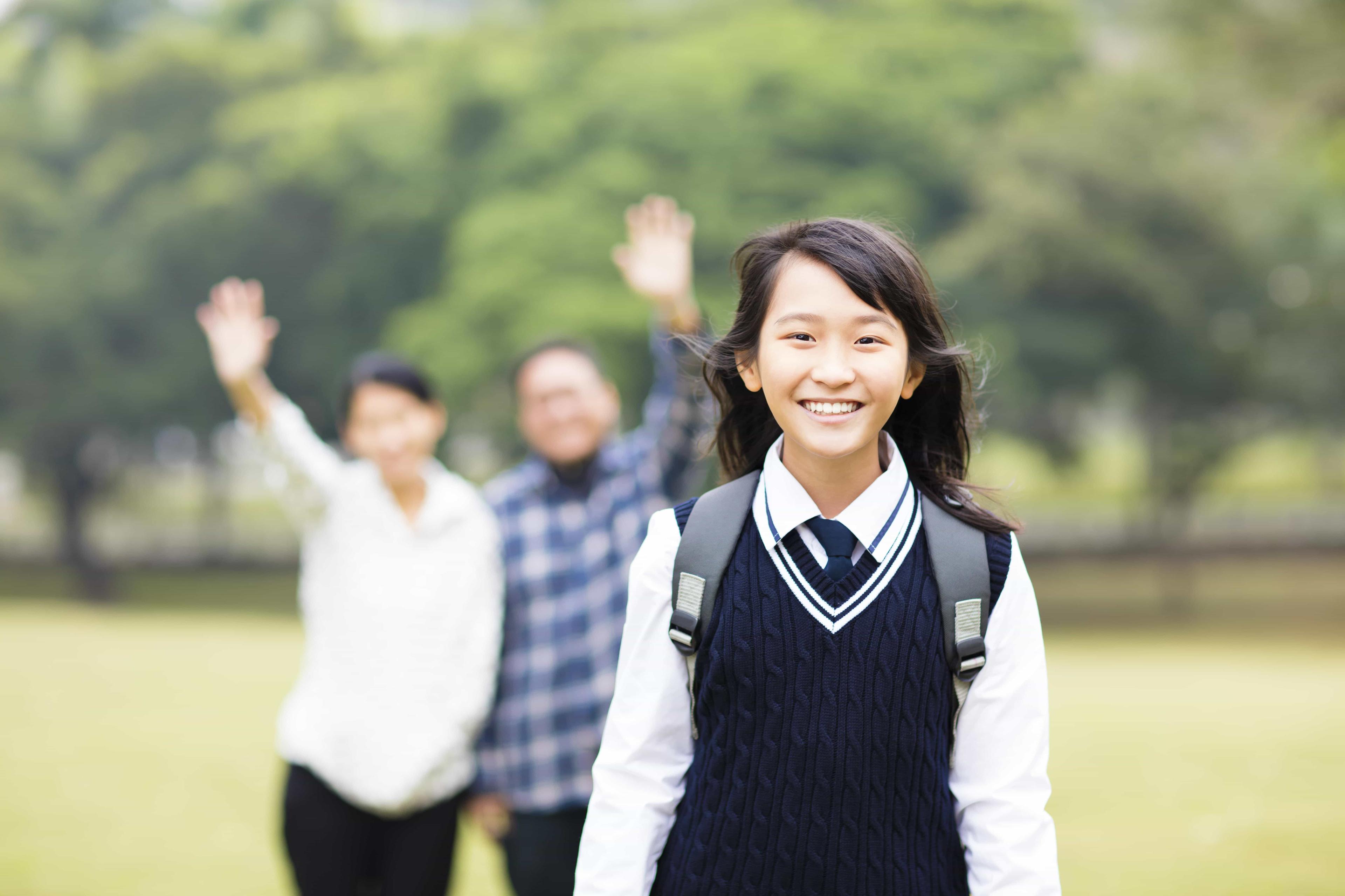 A young GCSE student smiles and her parents wave their hands in the background at their independent school.